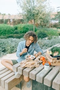 Wohnformen – Nachhaltiges & soziales Wohnen mit Symbiovita (e.V.) 8 Wohnformen – Nachhaltiges & soziales Wohnen mit Symbiovita (e.V.) A young man sitting at a picnic table, enjoying a fresh meal surrounded by greenery.